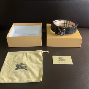 Svart skärp med silverdetalj - Snyggt svart bälte med klassiskt rutigt mönster på insidan och en stor silverfärgad 'B'-logga som spänne. Bältet är gjort i skinn och har exklusiv känsla, perfekt för att lyfta vilken outfit som helst.