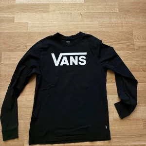 Svart långärmad tröja från Vans - Svart långärmad tröja från Vans med stort vitt logotryck på bröstet. Klassisk rund halsringning och mjukt material som känns skönt mot huden. Perfekt för dig som gillar streetwear och enkel stil. Passar folk mellan 160-175