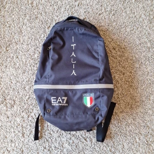 Mörkblå Emporio Armani EA7 x Italia ryggsäck - Snygg mörkblå Emporio Armani EA7 x Italia ryggsäck. Det är en collab mellan Emporio Armani EA7 och Italiens Olympiska-spelen lag. Ryggsäcken har använts många gånger och har lite slitningar (fråga gärna för fler bilder) men i stort sätt är skicket fortfarande väldigt bra. Skick: 7.5/10
