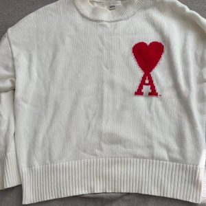 Vit stickad tröja från Ami - Vit stickad tröja från Ami med röd hjärtlogga på bröstet. Tröjan har rund halsringning och ribbade muddar vid ärmslut och nederkant. Perfekt för dig som gillar stilrena plagg med en twist.