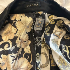 Versace blommig och randig skjorta - Unik skjorta från Versace med lyxigt blommigt och randigt mönster i svart, guld och beige. Sidenliknande material med detaljerade bokstäver och blommor, klassisk krage och guldfärgade knappar med Versace-logga. Perfekt för dig som vill sticka ut med en statement piece.