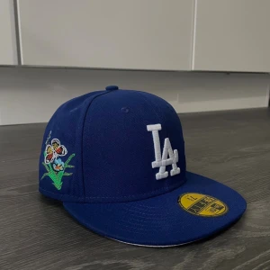 Blå LA New Era 59FIFTY keps med brodyr - Säljer en blå LA Dodgers 59FIFTY keps från New Era med broderade fjärilar, blommor och texten 'Felt' baktill. Kepsen har platt skärm, vitt LA-logo framtill och coola detaljer i flera färger. Perfekt för dig som vill sticka ut med din stil.