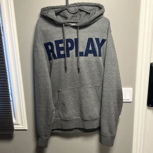 Grå hoodie från Replay med blå logga - Säljer en grå hoodie från Replay med stor blå logga framtill. Tröjan har huva med snörning och en klassisk känguruficka. Perfekt för dig som gillar streetwear och vill ha en bekväm och snygg tröja med tydlig branding. SKRIV VID FUNDERINGAR!!