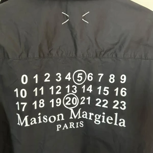 Maison Margiela svart skjorta med tryck - Svart skjorta från Maison Margiela med ikoniskt siffror-tryck på ryggen och bröstfickan. Skjortan har klassisk krage, långa ärmar och knäppning framtill. Materialet är bomull och designen har vita stygn-detaljer som ger en unik look. Använd men i fint skick. 