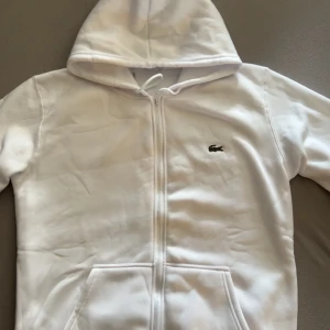 Lacoste zip hoodie - Snygg vit hoodie från Lacoste med hel dragkedja framtill och klassisk krokodillogga på bröstet. Tröjan har huva med snörning och två fickor framtill. Perfekt för en clean och sportig look.