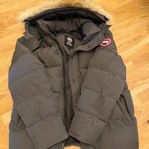 Mörkgrå dunjacka från Canada Goose - Säljer en mörkgrå dunjacka från Canada Goose med stor huva och äkta pälskant. Jackan har klassisk patch på ärmen, dragkedja och knappar framtill samt flera fickor. Perfekt för kalla vinterdagar och riktigt varm. Stilsäker och ikonisk modell äkta. 