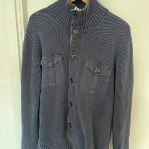 Mörkblå stickad kofta från Bondelid - Säljer en mörkblå stickad kofta från Bondelid med hög ribbad krage och knäppning framtill. Koftan har två bröstfickor med lock och knappar, samt ribbade muddar vid ärmslut och nederkant. Perfekt för lager-på-lager-stil under kyliga dagar.