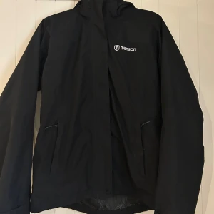 Svart regnjacka från Tenson med huva - Svart regnjacka från Tenson med huva och dragkedja. Jackan är vattentät, vindtät och samt andasfunktion. Två fickor framtill med dragkedja och diskret logga på bröstet och huvan. Perfekt för ruggiga dagar. Lite smuts på baksida luva