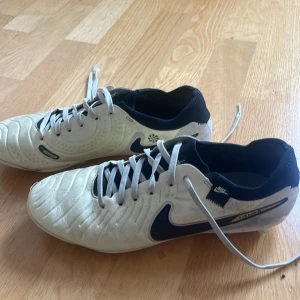 Nike Tiempo fotbollsskor vit/svart - Säljer ett par Nike Tiempo fotbollsskor i vitt och svart med guldiga detaljer. Skorna har snörning, rund tå och platt sula med dobbar för gräsplan. Ovandelen är i skinn och syntet, med klassisk Nike Swoosh på sidan och Tiempo-logga. Perfekta för dig som vill dominera på planen. Orginal pris 1200
