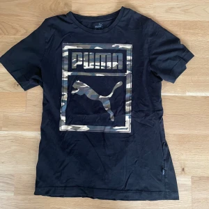 Svart Puma t-shirt med camo-tryck - Svart t-shirt från Puma med stort tryck på bröstet i camouflage-mönster och logotyp. Klassisk rund hals och korta ärmar. Materialet är mjuk bomull som känns skönt mot huden. Perfekt för dig som gillar sportig streetstyle och vill sticka ut med coolt print.