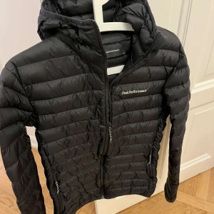 Svart dunjacka från Peak Performance🔥⚡️ - Säljer en snygg och varm svart dunjacka från Peak Performance. Perfekt för kyliga dagar med sin quiltade design och praktiska dragkedja. Jackan har en huva och är långärmad, vilket gör den idealisk för både höst och vinter. Passar lika bra till vardags som till utomhusaktiviteter.
