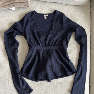 Långärmad Topp/Blus  - Marinblå blus. V-ringning där framme och knyte i ryggen, knyter går även att knyta framtill. Längre i armarna.