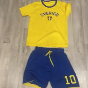 Säljer en gul och blå fotbollströja från H&M med texten 'Sverige' och ett litet emblem på bröstet. Tröjan är kortärmad och perfekt för unga fotbollsfans. Tillhörande blå shorts med nummer 10 ingår. Perfekt för att heja på Sverige i stil! 🇸🇪