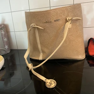 Beige handväska från Michael Kors - Säljer en snygg beige handväska från Michael Kors. Väskan har ett elegant mönster och är i syntetmaterial. Den har praktiska dragkedjor inuti för säker förvaring. Perfekt för både vardag och fest! Är fläckig inuti. 
