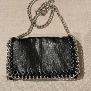 Svart skinnväska med silverkedja - Svart skinnväska med silverkedja som kan regleras till olika längder. Fint skick. Dragkedja för att stänga. 2 innerfack.  Mått: Bredd 20 cm Höjd 12 cm