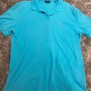 Ljusblå pikétröja från GANT - Snygg pikétröja från GANT med klassisk krage och knappdetalj. Broderad logotyp på bröstet ger en stilren touch. Perfekt för en avslappnad men ändå uppklädd look. Passar utmärkt för sommaren.