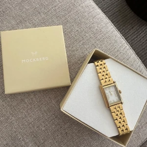 Guldfärgad klocka från Mockberg, Timeless Gold - Elegant guldfärgad klocka från Mockberg med rektangulär urtavla och romerska siffror. Armbandet är i metall med en klassisk länkdesign. Perfekt för en stilren look. Enbart använd 2 gånger då jag oftare använder andra klockor, den är för av i nyskick!