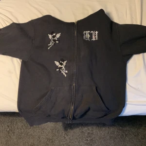 Bvaldi ”Guardians” - Snygg svart rhinestone hoodie från bvaldi och coola ängelmotiv på framsidan. Texten 'GUARDIANS' pryder ryggen i ett stilrent typsnitt. Perfekt för en avslappnad stil.