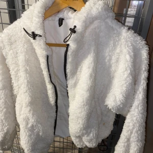 Vit teddyjacka med huva - Mysig vit teddyjacka från Boohoo med huva och dragkedja framtill. Perfekt för kyliga dagar när du vill hålla dig varm och stilren. Jackan har en fluffig textur och är helfodrad för extra komfort.