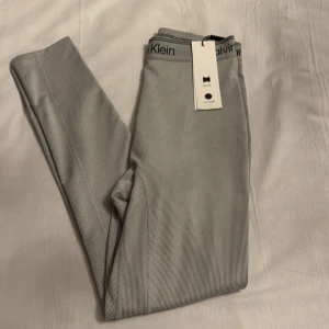 Grå ribbade leggings från Calvin Klein - Snygga grå ribbade leggings från Calvin Klein med elastisk midja och logotypdetaljer. Perfekta för en sportig look eller avslappnad stil. Superbekväma och stilrena!