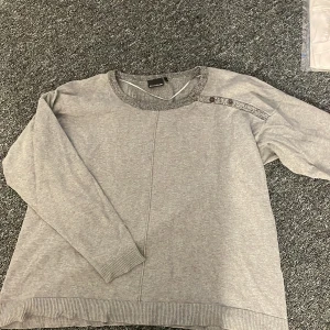 Grå tröja med påfågelmönster - Säljer en grå tröja med ett unikt påfågelmönster på baksidan. Tröjan har långa ärmar och ribbade kanter vid hals, ärmslut och nederkant. Perfekt för en avslappnad stil.