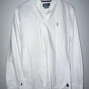 Vit skjorta från Polo Ralph Lauren - Snygg vit skjorta från Polo Ralph Lauren i slim fit. Skjortan har långa ärmar och klassiska knappar framtill. Den är broderad med den ikoniska logotypen på bröstet. Perfekt för en stilren look.