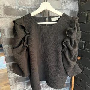 Svart blus med volangärmar  - Snygg svart blus från med volangdetaljer på ärmarna. Perfekt för en stilren look. Blusen har en rund halsringning och knäppning i nacken.