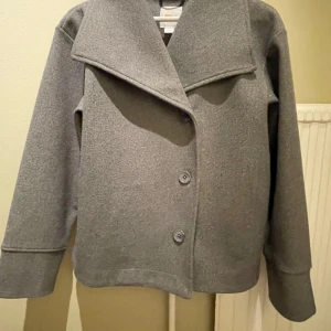 Grå kappa med bred krage - Snygg grå kappa med bred krage och knappar framtill. Perfekt för kyligare dagar med sin stilrena design och långa ärmar. Är i storlek 158/164 men passar mig som brukar ha S/XS