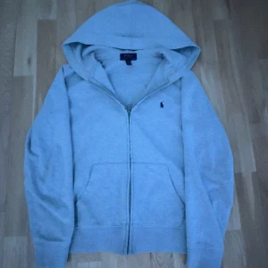 Grå hoodie från Polo Ralph Lauren - Säljer en stilren grå hoodie från Polo Ralph Lauren med dragkedja och klassisk logga på bröstet. Perfekt för en avslappnad look med praktiska fickor framtill och en bekväm huva.