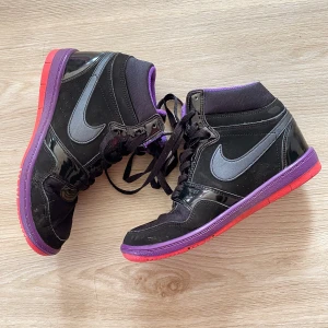 Nike  Force sky high i svart och lila - Snygga Nike  Force sky high sneakers i svart med lila och röda detaljer. Skorna har en hög modell med snörning och en iögonfallande röd sula. Perfekta för att ge din outfit en cool touch.