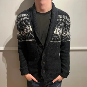 Svart kofta med mönster - Snygg svart kofta med ett stilrent vitt och grått mönster över axlarna. Koftan har knappar framtill och ribbade muddar vid ärmslut. Perfekt för kyligare dagar. SIZE S MEN PASSAR M. FÖR DÖM MED MYCKET MUSKLER SKULLE DEN VARA FÖR LITRN TYVÄRR🔥🔥🔥🙋