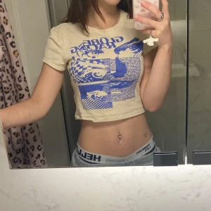 Beige croppad top   - Säljer en beige croppad t-shirt med ett coolt blått tryck från HM. T-shirten har korta ärmar och en rund halsringning. Perfekt för en avslappnad stil.