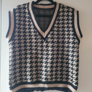 Svart och beige stickad väst, Hundtand, M/L - Snygg stickad väst i svart och beige med ett klassiskt hundtandsmönster. Västen har en v-ringad design och ribbade kanter som ger en stilren look. Perfekt för lager-på-lager under kyligare dagar. Stl M/L rymlig boxig modell. Sparsamt använd, väldigt välhållen!