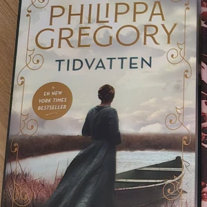 Tidvatten - Boken 'Tidvatten' av Philippa Gregory är en New York Times bestseller. Omslaget visar en kvinna i en lång klänning vid en flod med en båt i bakgrunden. Perfekt för dig som älskar historiska romaner och dramatik.