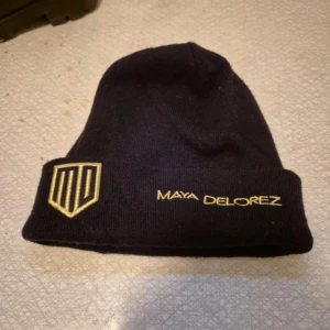 Svart mössa från Maya Delorez - Snygg svart mössa från Maya Delorez med broderad logotyp och text i guld. Perfekt för att hålla värmen under kyliga dagar. Mössan är använd ca 5 gånger, och köptes för ca 400kr. Pris kan sänkas vid snabbaffär.