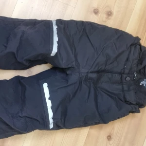 Svarta byxortäckbyxor / skidbyxor från Kaxs - Svarta byxor slidbyxor från Kaxs med reflexdetaljer, dock är reflexerna slitna. Byxorna har en justerbar midja och stängs med knapp och dragkedja. Perfekta för utomhusaktiviteter under vintern. Använda en vinter av ett barn