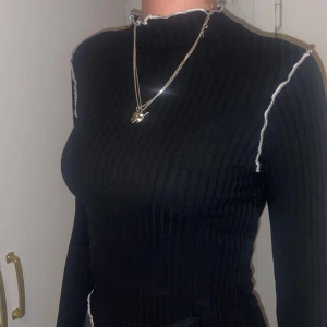 Svart ribbad tröja med vita detaljer - Snygg svart ribbad tröja med vita kontrastsömmar. Tröjan har en hög krage och långa ärmar, perfekt för en stilren look. Passar bra till både jeans och kjol.