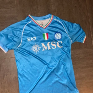 Blå Napoli fotbollströja med nummer 77 - Snygg blå Napoli fotbollströja från EA7 med korta ärmar och nummer 77 på ryggen. Tröjan har klubbens emblem och sponsorloggor på framsidan samt italienska flaggans färger vid kragen.