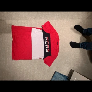 Röd och vit t-shirt från Michael Kors - Snygg röd och vit t-shirt från Michael Kors med stor logga på bröstet. T-shirten har en klassisk passform och korta ärmar, perfekt för en avslappnad stil.