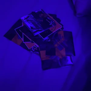 Klibbor vet ej hur många  - En hög med färgglada kort eller klistermärken i olika mönster och färger ligger på ett underlag belyst av blått ljus. Det är svårt att urskilja exakta motiv eller detaljer på korten på grund av belysningen.