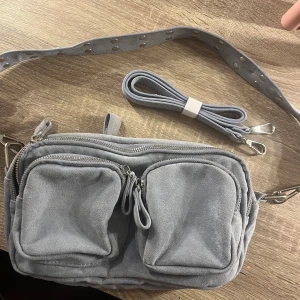Blå axelväska med dragkedjor - Snygg blå/grå axelväska i mocka med flera praktiska fack och dragkedjor. Väskan har en axelrem med nitar och en extra rem för variation. Den är rymlig och man får plats med mycket, dock har den tyvärr inte kommit så mycket till användning. Från Gina⭐️