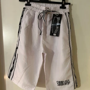 Vita shorts från Urban Style - Snygga vita shorts från Urban Style med svart broderad textdetalj. De har elastisk midja med snörning och svarta ränder längs sidorna. Perfekta för en avslappnad stil.