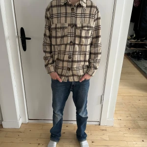 Snygg Gruntskjorta till barn - Säljer en beige-rutig overshirt från Grunt i storlek M. Skjortan är tillverkad i ett mjukt och varmt fleecematerial och har två bröstfickor med knappar. Perfekt som en lätt jacka eller som ett extra lager under kyliga dagar. Väldigt lite använd.  Personen på bilderna är 160 cm lång, 12 år gammal och har en smal/normal kroppsbyggnad.  Skick: Bra begagnat skick med minimala tecken på användning.  Nypris: 500 SEK