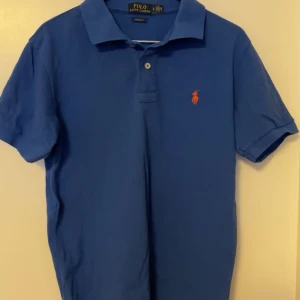 Blå pikétröja från Ralph Lauren - Säljer en klassisk blå pikétröja från Ralph lauren. Tröjan har korta ärmar och en stilren krage med knappar. Perfekt för en avslappnad men stilfull look.