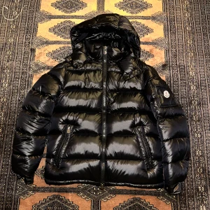Svart pufferjacka från Moncler - Snygg svart pufferjacka från Moncler med huva och dragkedja. Jackan har en glansig finish och är perfekt för kyliga dagar. Den har långa ärmar och praktiska fickor med knappar.