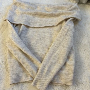Beige stickad offshoulder-tröja - Mysig beige stickad tröja med offshoulder-design. Perfekt för kyliga dagar med sin mjuka och bekväma passform. Tröjan har långa ärmar och ribbade detaljer vid ärmslut och nederkant.