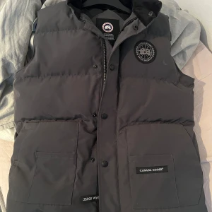 Svart dunväst från Canada Goose - Säljer en svart dunväst från Canada Goose med dragkedja och knappar. Västen har en broderad logotyp på bröstet och praktiska fickor framtill. Perfekt för kyliga dagar.