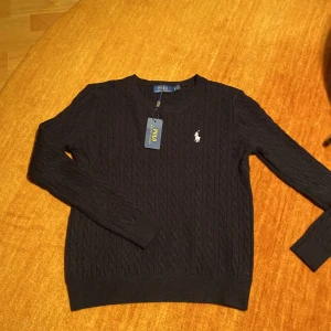 Svart kabelstickad tröja från Ralph Lauren - Säljer en stilren svart kabelstickad tröja från Ralph Lauren med det klassiska logotypbroderiet på bröstet. Tröjan har en rund halsringning och långa ärmar, perfekt för en elegant och avslappnad look. Fick denna tröja i julklapp men satt inte direkt som jag ville därför säljs den nu.