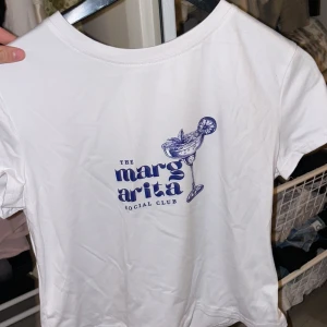 Vit t-shirt med Margarita-tryck - Snygg vit t-shirt med ett coolt blått tryck av en margarita och texten 'The Margarita Social Club'. Perfekt för en avslappnad stil. Ej stryken, därför är den rynkig. 