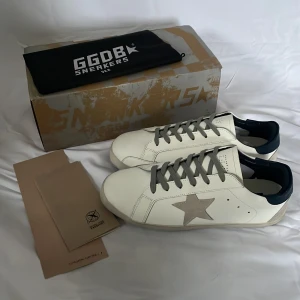 Vit GGDB skor - Snygga vita sneakers från Golden Goose med en grå stjärndetalj på sidan. Skorna har grå snörning och en svart häl. Perfekta för en stilren look.